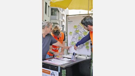 Drei Personen unter einem gelben Sonnenschirm betrachten gemeinsam einen großen Lageplan an einem Info-Stand. Die Person links trägt eine dunkle Brille und graues Shirt, die mittlere Person trägt eine orange Warnweste und die rechte Person trägt einen dunklen Pullover mit oranger Weste. Auf dem Tisch vor ihnen liegen Unterlagen und ein Glas mit Stiften. Der Plan zeigt einen Stadtplan mit grünen Notiz-Zetteln. Im Hintergrund mehrstöckige Wohnhäuser. Die Szene wirkt wie ein Bürgerbeteiligungsprozess."
