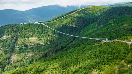 Eine weite Landschaftsaufnahme zeigt eine lange, filigrane Hängebrücke, die sich über ein bewaldetes Tal spannt, die Konstruktion verbindet zwei Höhenzüge und ist deutlich in die Topografie eingebettet, wodurch das Bild Ingenieurbau als landschaftsprägendes Element zeigt und für Erschließung, Erlebnis und respektvollen Umgang mit Naturraum steht.