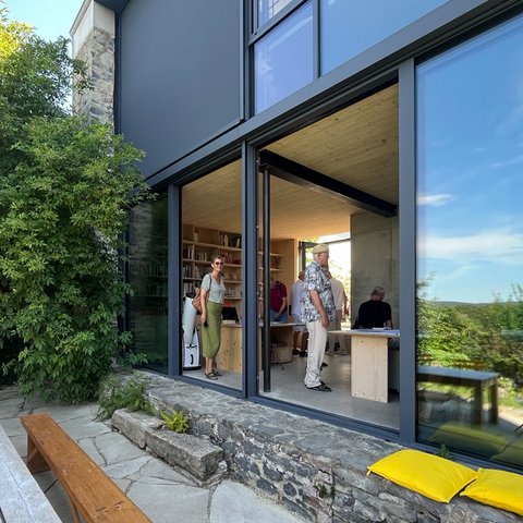 „Blick über eine sonnige Terrasse mit Holztisch auf ein modernes Haus mit raumhoher Glasfront. Durch die offenen Fenster sieht man Personen im Innenraum, während sich in den Scheiben die grüne Landschaft spiegelt.“