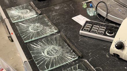 Vier kleine Verbundglasscheiben mit sternförmigen Rissen nach Schlagprüfung auf Labortisch; daneben Handschuh und Messgeräte.