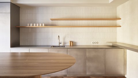 Minimalistische Küche mit grauen Fronten, heller Fliesenrückwand und offenen Holzregalen. Im Vordergrund steht ein runder Holztisch auf einem Fischgrätparkett.