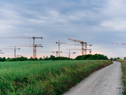 Mehrere Baukräne stehen hinter einer grünen Wiese; ein schmaler Weg führt durch die Landschaft, im Hintergrund Baustellen und niedrige Bebauung unter bewölktem Himmel.