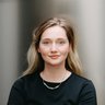 Halbnahes Farbfoto einer jungen Frau: hellblondes, schulterlanges Haar, dezentes Make‑up und leichtes Lächeln, sie trägt ein schwarzes Oberteil und eine kurze silberne Kette; frontal aufgenommen vor einem neutralen, leicht reflektierenden Hintergrund.