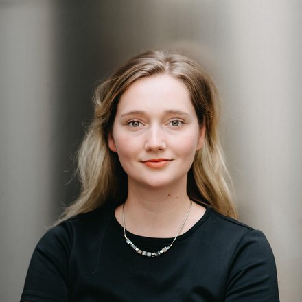 Halbnahes Farbfoto einer jungen Frau: hellblondes, schulterlanges Haar, dezentes Make‑up und leichtes Lächeln, sie trägt ein schwarzes Oberteil und eine kurze silberne Kette; frontal aufgenommen vor einem neutralen, leicht reflektierenden Hintergrund.