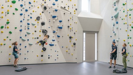 Weiße Kletterwand in moderner Kletterhalle, mehrere Menschen mit Klettergurten