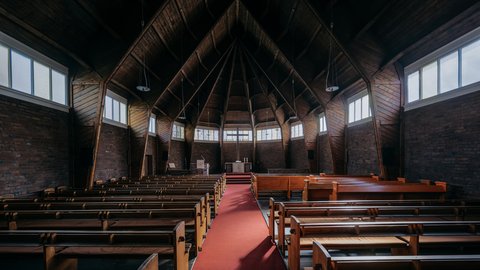 Kirchenschiff mit dunkler Holzkonstruktion, symmetrisch angeordneten Sitzbänken und zentralem Gang, der zum Altar im hinteren Bereich führt.