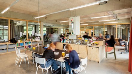 Großzügiger, offener Innenraum mit Möbeln und Kücheninsel: helle Mehrzweckfläche mit großen Tischen, Stühlen und Holzschränken, durchsichtige Trennwände zu angrenzenden Räumen; mehrere Personen sitzen oder gehen herum, Bewegungsunschärfen zeigen lebhafte Alltagsszene von Lernen oder gemeinsamer Arbeit.