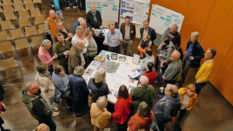 "Luftaufnahme einer Gruppe von etwa 30 Personen, die im Kreis um einen zentralen Tisch stehen. Auf dem Tisch liegen Pläne und Dokumente. Mehrere Flip-Charts mit handschriftlichen Notizen und Diagrammen sind im Hintergrund sichtbar. Graue Stühle stehen aufgereiht im oberen Bereich des Raumes. Der Boden ist dunkel. Die Teilnehmer tragen verschiedenfarbige Kleidung und hören einem Sprecher zu, der in der Mitte der Gruppe steht. Dies scheint ein professioneller Workshop oder Planungstreffen zu sein."