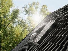 „Ein modernes Dachfenster auf einem dunklen Ziegeldach, angestrahlt von hellem Sonnenlicht vor dem Hintergrund grüner Baumkronen.“