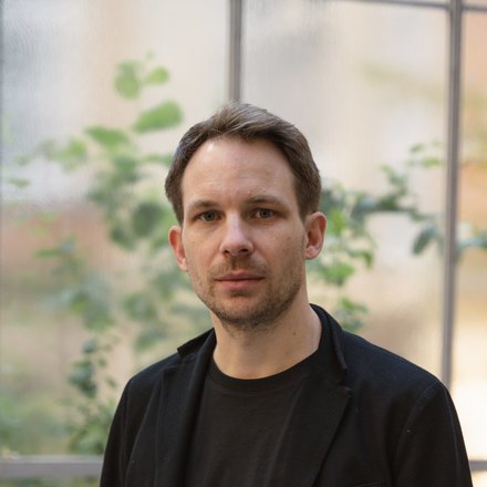 Person in schwarzer Kleidung steht vor milchigen Glasfenstern, dahinter unscharfe grüne Pflanzen.