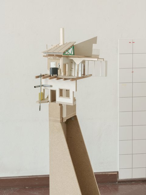  Eine Frontalansicht eines architektonischen Architekturmodells, das auf einem hohen Sockel aus Spanplatten steht. Das Modell zeigt eine detaillierte Schnittdarstellung eines mehrstöckigen Gebäudes mit weißem Verputz, Holzskelettkonstruktion und verschiedenen Dachformen. Zu den Details gehören Fensterrahmen aus Holz, ein grünes Fachwerkelement im Dachstuhl und ein schwarzer Kamin. Der Sockel ist schmal und ragt weit auf. Das Bild ist vor einem neutralen weißen Hintergrund mit einem gefliesten Bereich auf der rechten Seite aufgenommen.