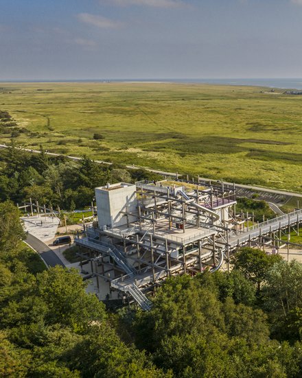 Luftaufnahme einer weiten, grünen Marschlandschaft an der Küste mit Wasserlauf und Sandbänken am Horizont. Im Vordergrund steht ein mehrstöckiges, offenes Stahlbauwerk mit Rampen und Röhrenrutschen, verbunden mit einem langen erhöhten Steg; daneben verlaufen Wege mit Radfahrenden zwischen Bäumen.