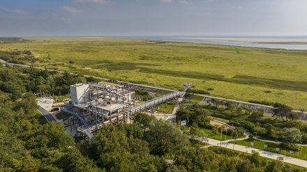 Luftaufnahme einer weiten, grünen Marschlandschaft an der Küste mit Wasserlauf und Sandbänken am Horizont. Im Vordergrund steht ein mehrstöckiges, offenes Stahlbauwerk mit Rampen und Röhrenrutschen, verbunden mit einem langen erhöhten Steg; daneben verlaufen Wege mit Radfahrenden zwischen Bäumen.