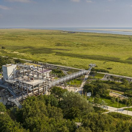 Luftaufnahme einer weiten, grünen Marschlandschaft an der Küste mit Wasserlauf und Sandbänken am Horizont. Im Vordergrund steht ein mehrstöckiges, offenes Stahlbauwerk mit Rampen und Röhrenrutschen, verbunden mit einem langen erhöhten Steg; daneben verlaufen Wege mit Radfahrenden zwischen Bäumen.