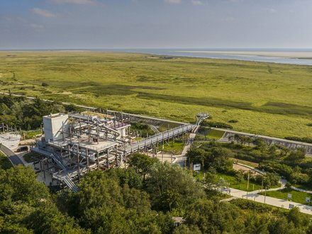 Luftaufnahme einer weiten, grünen Marschlandschaft an der Küste mit Wasserlauf und Sandbänken am Horizont. Im Vordergrund steht ein mehrstöckiges, offenes Stahlbauwerk mit Rampen und Röhrenrutschen, verbunden mit einem langen erhöhten Steg; daneben verlaufen Wege mit Radfahrenden zwischen Bäumen.