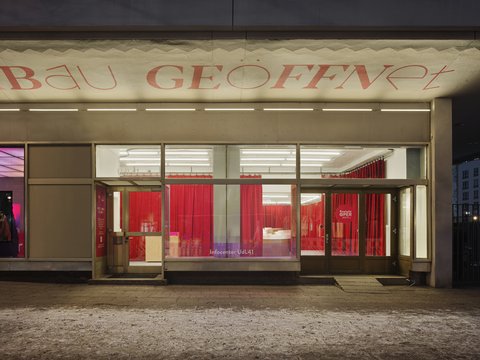 "Nachtaufnahme eines hell erleuchteten Geschäftsgebäudes. Der überdachte Eingangsbereich zeigt in roten Buchstaben den Schriftzug 'BAU GEÖFFNET'. Durch die großen Schaufenster blickt man in einen Ausstellungsraum, der von leuchtend roten Samtvorhängen dominiert wird. Am rechten Fensterrand klebt ein runder roter Aufkleber mit der Aufschrift 'KOMISCHE OPER BERLIN'. Der Gehweg davor ist mit etwas Schnee bedeckt."