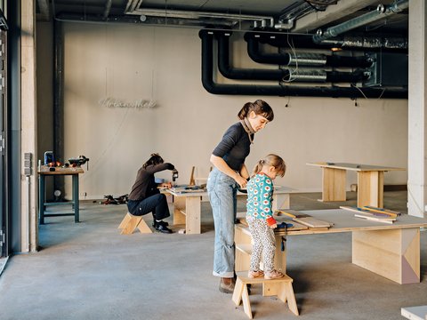 Frau hilft Kind beim Arbeiten an einem Holztisch in einer Werkstatt, im Hintergrund sitzt eine weitere Person auf einem Hocker und arbeitet an einem Tisch