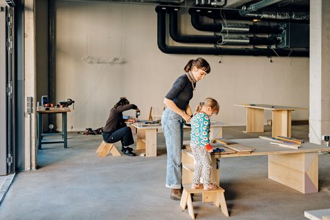 Frau hilft Kind beim Arbeiten an einem Holztisch in einer Werkstatt, im Hintergrund sitzt eine weitere Person auf einem Hocker und arbeitet an einem Tisch