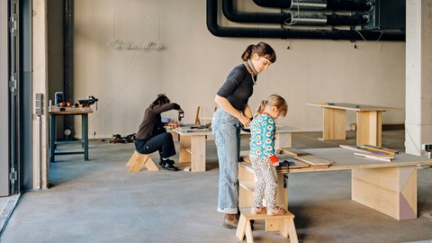 Frau hilft Kind beim Arbeiten an einem Holztisch in einer Werkstatt, im Hintergrund sitzt eine weitere Person auf einem Hocker und arbeitet an einem Tisch