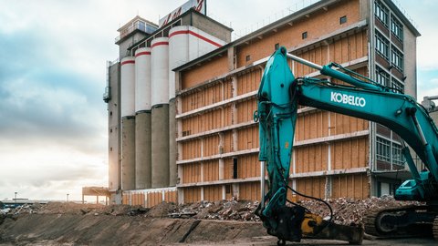 Braunes Gebäude mit vier Säulen links und einem grünen Bagger im Vordergrund