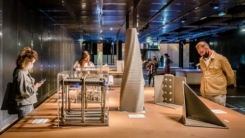 "Innenraum einer Ausstellung mit Fokus auf Bau- und Konstruktionsmaterialien. Auf einem langen Tisch stehen verschiedene Modelle, darunter ein Würfel aus Stahlbewehrung in einer transparenten Vitrine und ein hohes, spiralförmiges Objekt. Drei Besucher betrachten die Exponate aus nächster Nähe. Der Raum ist modern gestaltet mit dunklen Wänden und gerichteter Beleuchtung."