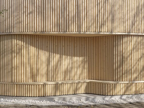 "Nahaufnahme einer vertikalen Holzlamellenfassade aus hellen Holzlamellen. Drei horizontale Betongürtel unterbrechen die senkrechte Holzstruktur. Sonnenlicht erzeugt längliche Schattenmuster auf der Oberfläche. Grauer Kiesstreifen am unteren Rand."