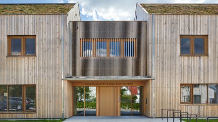 Eingangsfassade der Kindertagesstätte in Holzbauweise; über der Tür steht „Kindertagesstätte Burg Wichtelstein“, begrüntes Dach sichtbar.