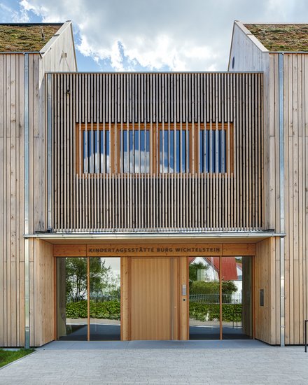 Eingangsfassade der Kindertagesstätte in Holzbauweise; über der Tür steht „Kindertagesstätte Burg Wichtelstein“, begrüntes Dach sichtbar.