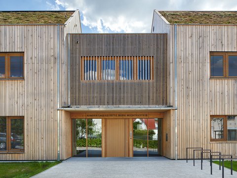 Eingangsfassade der Kindertagesstätte in Holzbauweise; über der Tür steht „Kindertagesstätte Burg Wichtelstein“, begrüntes Dach sichtbar.