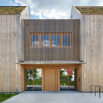 Eingangsfassade der Kindertagesstätte in Holzbauweise; über der Tür steht „Kindertagesstätte Burg Wichtelstein“, begrüntes Dach sichtbar.