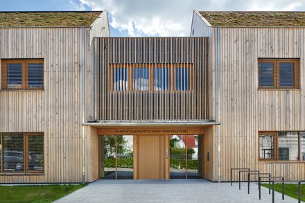 Eingangsfassade der Kindertagesstätte in Holzbauweise; über der Tür steht „Kindertagesstätte Burg Wichtelstein“, begrüntes Dach sichtbar.