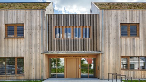 Eingangsfassade der Kindertagesstätte in Holzbauweise; über der Tür steht „Kindertagesstätte Burg Wichtelstein“, begrüntes Dach sichtbar.