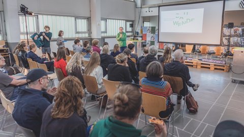 Person steht vor Publikum in einem hellen Raum mit großen Fenstern und präsentiert vor einer Leinwand