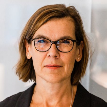 Farbfoto: Nahaufnahme einer Person mit braunen, schulterlangen Haaren und dunkler Brille; neutraler Blick; heller, unscharfer Bürohintergrund.