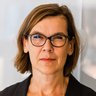 Farbfoto: Nahaufnahme einer Person mit braunen, schulterlangen Haaren und dunkler Brille; neutraler Blick; heller, unscharfer Bürohintergrund.