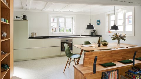 "Großzügige, helle Wohnküche mit offener Raumaufteilung. Links moderne Küche mit hellgrünen Schränken, schwarzer Arbeitsplatte und Edelstahlgeräten. In der Mitte großer Holzesstisch mit grünen Stühlen. Rechts Holzregal mit Büchern. Decke mit Balkenoptik, mehrere helle Fenster sorgen für natürliche Beleuchtung. Schwarze Pendelleuchten über dem Tisch."