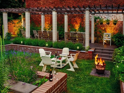 Gemütlicher, beleuchteter Garten mit Sitzgruppe, Feuerkorb und Pergola vor Backsteinwand.