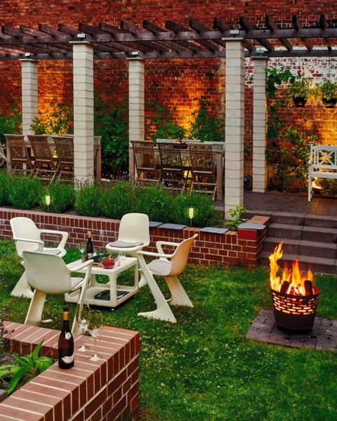 Gemütlicher, beleuchteter Garten mit Sitzgruppe, Feuerkorb und Pergola vor Backsteinwand.