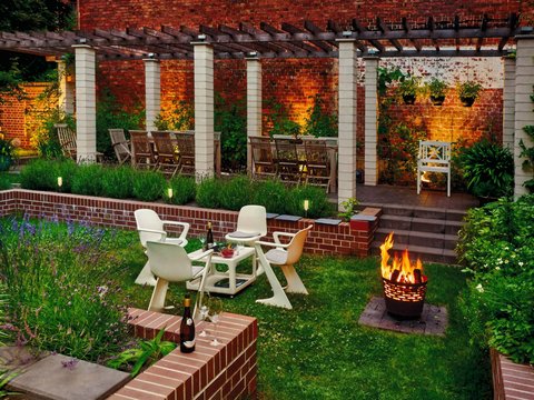 Gemütlicher, beleuchteter Garten mit Sitzgruppe, Feuerkorb und Pergola vor Backsteinwand.