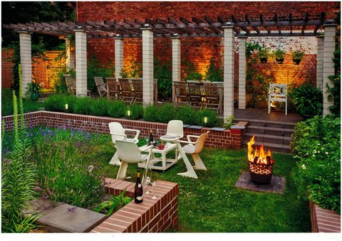 Gemütlicher, beleuchteter Garten mit Sitzgruppe, Feuerkorb und Pergola vor Backsteinwand.