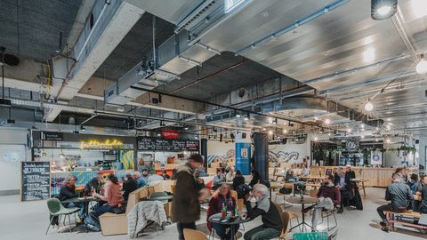 Belebte Markthalle mit Tischen, Sitzgelegenheiten und vielen Menschen beim Essen und Unterhalten.