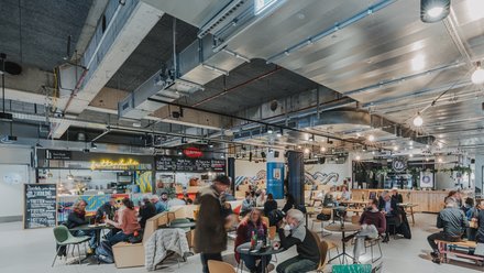 Belebte Markthalle mit Tischen, Sitzgelegenheiten und vielen Menschen beim Essen und Unterhalten.