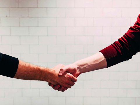 Zwei Personen geben sich die Hand zum Händedruck; zu sehen sind nur Unterarme und Hände, eine Person trägt einen roten, die andere einen schwarzen Ärmel, im Hintergrund eine helle, geflieste Wand.