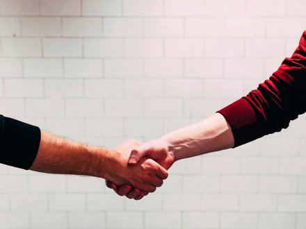 Zwei Personen geben sich die Hand zum Händedruck; zu sehen sind nur Unterarme und Hände, eine Person trägt einen roten, die andere einen schwarzen Ärmel, im Hintergrund eine helle, geflieste Wand.
