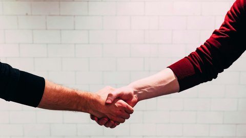 Zwei Personen geben sich die Hand zum Händedruck; zu sehen sind nur Unterarme und Hände, eine Person trägt einen roten, die andere einen schwarzen Ärmel, im Hintergrund eine helle, geflieste Wand.