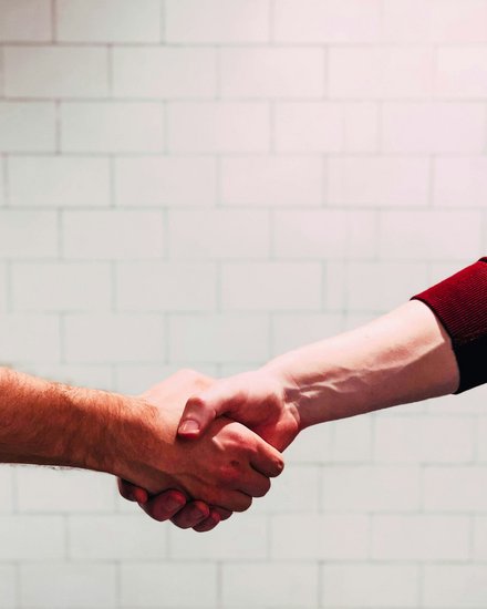 Zwei Personen geben sich die Hand zum Händedruck; zu sehen sind nur Unterarme und Hände, eine Person trägt einen roten, die andere einen schwarzen Ärmel, im Hintergrund eine helle, geflieste Wand.