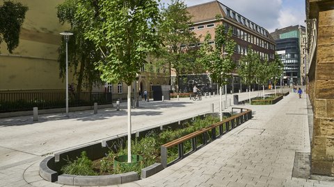 "Luftaufnahme eines modernen, gut ausgestatteten Stadtplatzes an einem sonnigen Tag. Der Platz ist mit hellem Naturstein gepflastert. Im Vordergrund verlaufen mehrere lange, rechteckige Hochbeete mit dunkelgrünem Metallgeländer und Bepflanzung. Weiße Metallstraßenlaternen stehen verteilt auf dem Platz. Am Rand des Platzes sind historische rote Backsteingebäude verschiedener Größen sichtbar. Auf dem Platz sind mehrere Menschen zu sehen, darunter ein Fahrradfahrer. Die Szene wirkt offen, modern gestaltet und öffentlich zugänglich."
