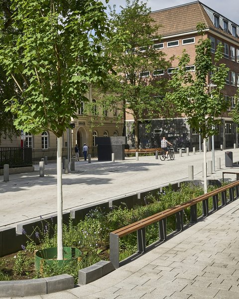 "Luftaufnahme eines modernen, gut ausgestatteten Stadtplatzes an einem sonnigen Tag. Der Platz ist mit hellem Naturstein gepflastert. Im Vordergrund verlaufen mehrere lange, rechteckige Hochbeete mit dunkelgrünem Metallgeländer und Bepflanzung. Weiße Metallstraßenlaternen stehen verteilt auf dem Platz. Am Rand des Platzes sind historische rote Backsteingebäude verschiedener Größen sichtbar. Auf dem Platz sind mehrere Menschen zu sehen, darunter ein Fahrradfahrer. Die Szene wirkt offen, modern gestaltet und öffentlich zugänglich."