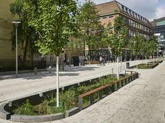 "Luftaufnahme eines modernen, gut ausgestatteten Stadtplatzes an einem sonnigen Tag. Der Platz ist mit hellem Naturstein gepflastert. Im Vordergrund verlaufen mehrere lange, rechteckige Hochbeete mit dunkelgrünem Metallgeländer und Bepflanzung. Weiße Metallstraßenlaternen stehen verteilt auf dem Platz. Am Rand des Platzes sind historische rote Backsteingebäude verschiedener Größen sichtbar. Auf dem Platz sind mehrere Menschen zu sehen, darunter ein Fahrradfahrer. Die Szene wirkt offen, modern gestaltet und öffentlich zugänglich."