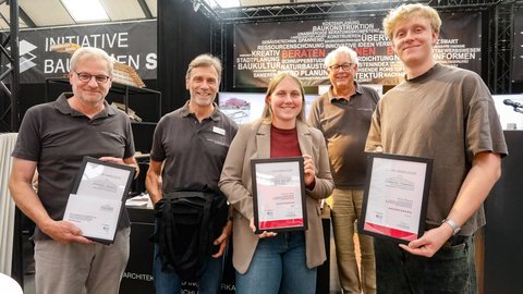 Kleine Gruppe mit Auszeichnungen posiert vor einer Ausstellungswand zum Thema Bauen und Planen.